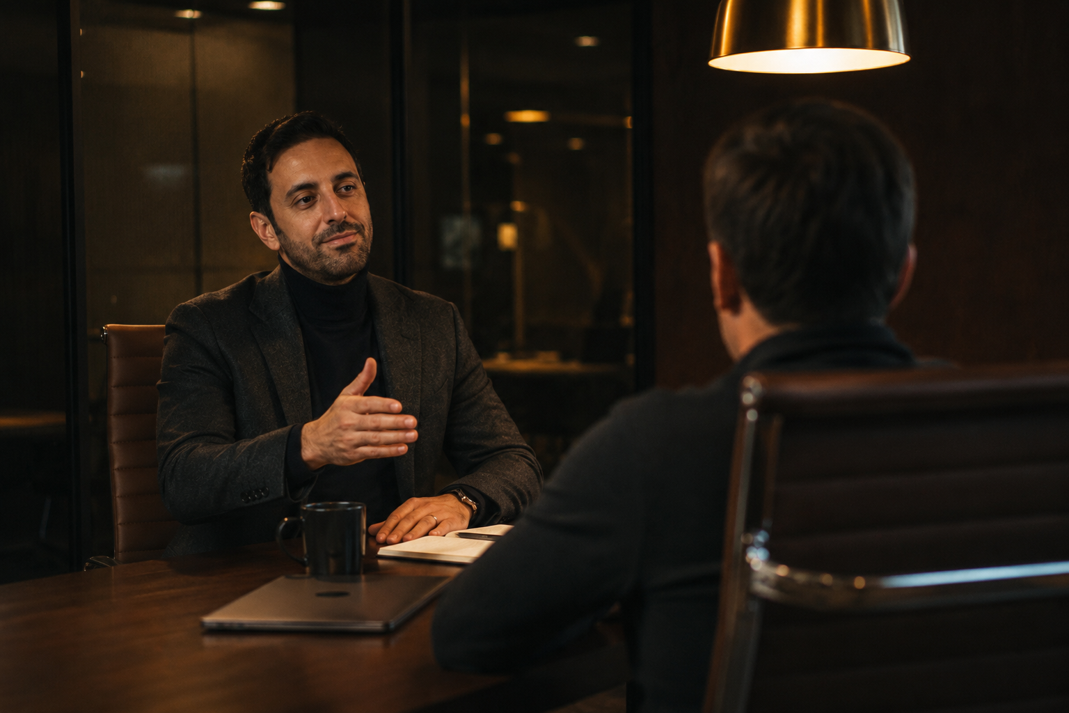Yusuf gesturing across a dark walnut conference table to a founder, two leather chairs, single brass pendant lamp casting warm pool of light, smoked glass wall behind